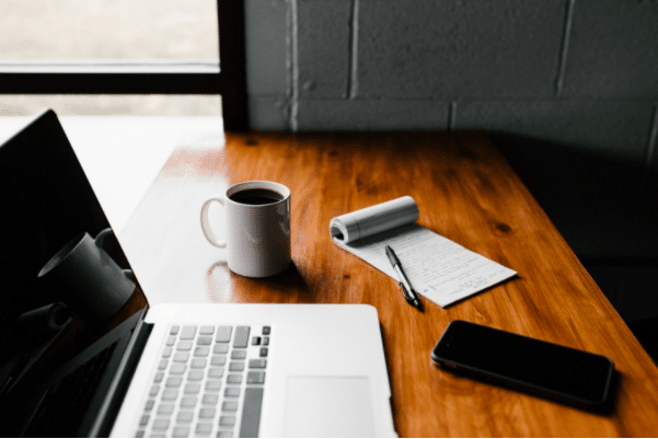 On a wooden desk sits an open laptop, a phone, a mug of coffee and a notepad and pen. The notepad appears to have lots of writing on it.