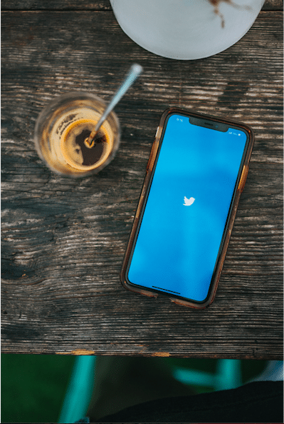On a brown wooden table lies an iphone with the Twitter opening page on the screen. It is a bright blue with a white bird icon in the middle. Next to it is an almost empty glass of coffee with a metal spoon in it.