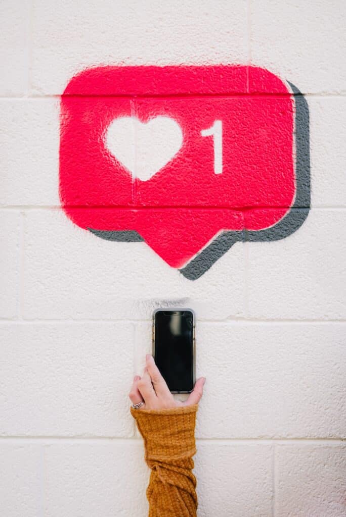 A hand holds up a smart phone against a white wall. Above, is a pink speech bubble with a white love heart and the number 1 next to each other, representing a reaction to a social media post.