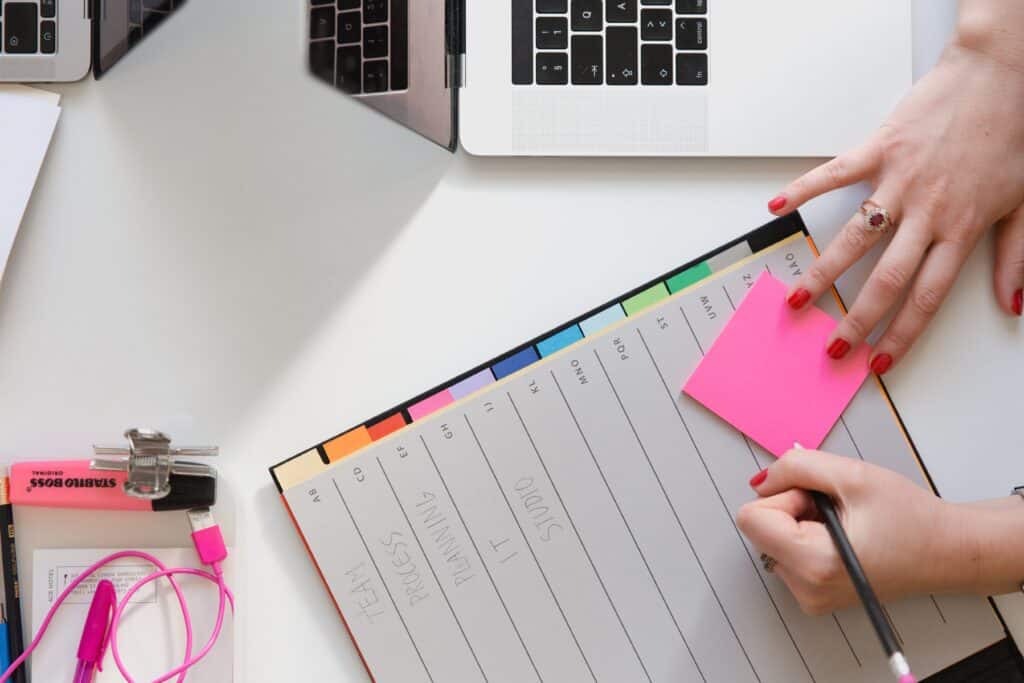 On a white table there is a laptop, a notepad and a highlighter. A person's hand with red painted fingernails and a ring on, write in the notepad.