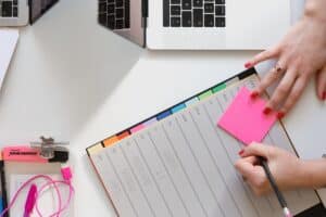 On a white table there is a laptop, a notepad and a highlighter. A person's hand with red painted fingernails and a ring on, write in the notepad.