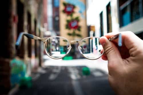 A persons hand holds a pair of glasses out in front of them on a street. The area around is blurred but the lenses are in focus.