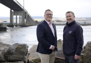Two men standing on rocky ground near a large bridge, with a river in the background. One man wears a navy blazer and pink shirt with glasses, the other wears a dark fleece with a Scottish Water logo. They are both smiling and facing the camera, with an industrial bridge structure and urban landscape behind them. Ross Mclory and Ian Ferguson