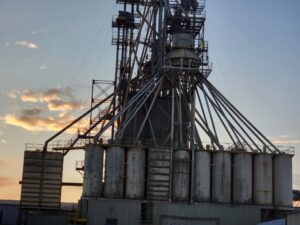 Industrial grain silos and feed mill structure at sunset. The large metal storage bins are supported by steel framework with elevated conveyor systems and loading equipment. The silhouette of the facility is dramatically backlit by a colorful dusk sky with scattered clouds.