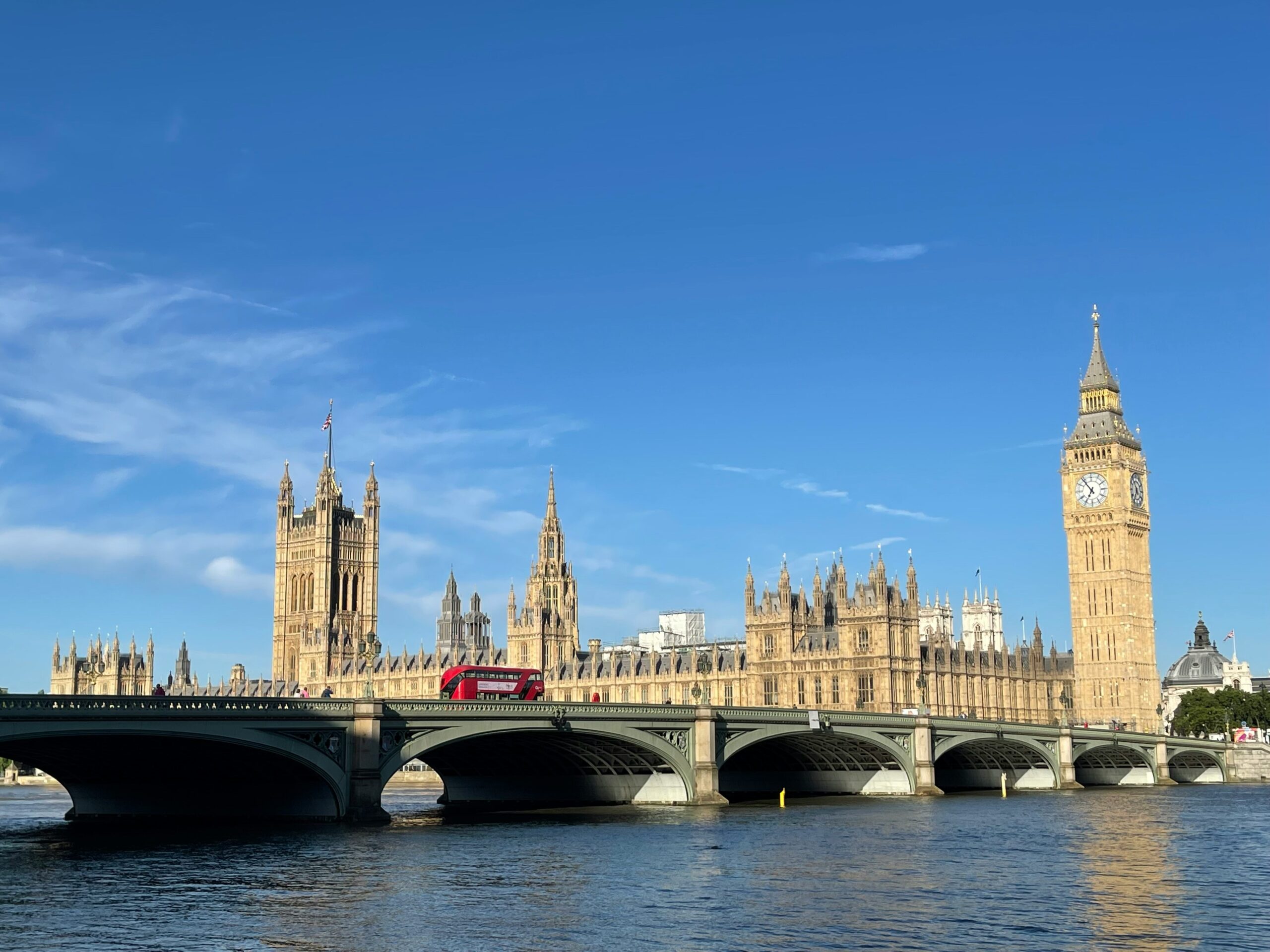 A stunning view of the Houses of Parliament and Elizabeth Tower (Big Ben) in London on a clear sunny day. Westminster Bridge spans across the River Thames in the foreground with a red double-decker bus visible crossing the bridge. The Gothic architecture of the Parliament buildings stands majestically against a bright blue sky.