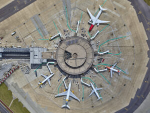 This aerial drone image shows a circular airport terminal with approximately ten aircraft docked at gates arranged in a radial pattern. The terminal has a distinctive circular design with a central hub and passenger boarding bridges (jetways) extending outward like spokes of a wheel. Various commercial aircraft in different liveries (including white with red tail, orange, and yellow accents) are connected to these jetways. The concrete apron surrounds the terminal, with clear taxiway markings visible. Ground service vehicles and equipment can be seen near the aircraft. The photo captures the symmetrical layout of the terminal from directly above, showing the efficient use of space in this hub-and-spoke airport design. Runways and additional airport infrastructure are partially visible at the edges of the frame.