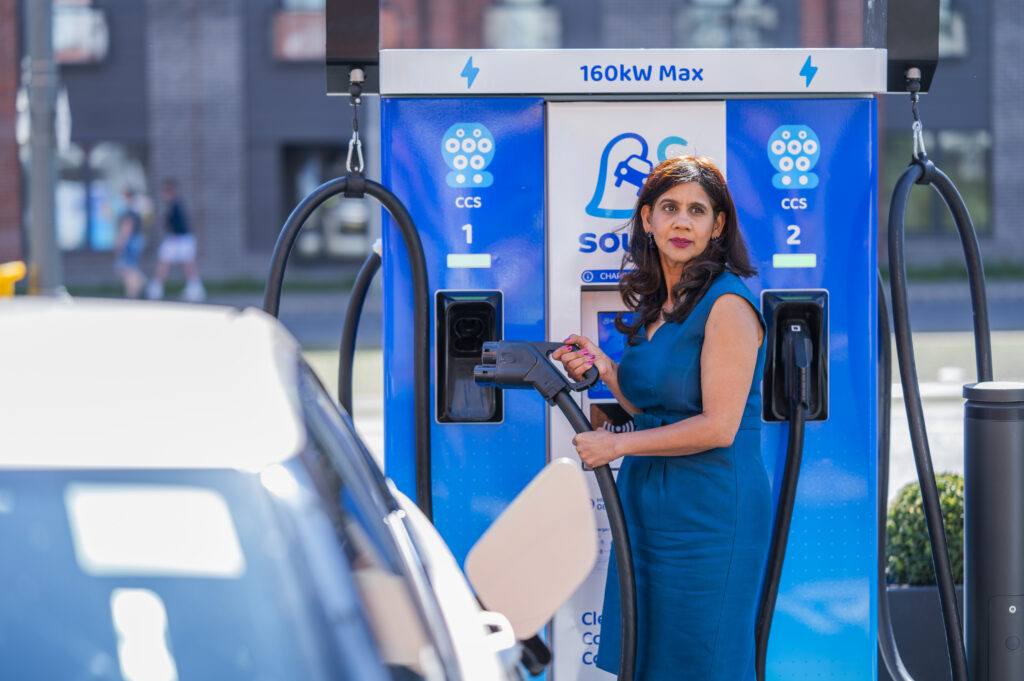 Deepa Chandrasekaran beside a blue EV at the new Source charging hub in Edinburgh, charging the car and holding the plug
