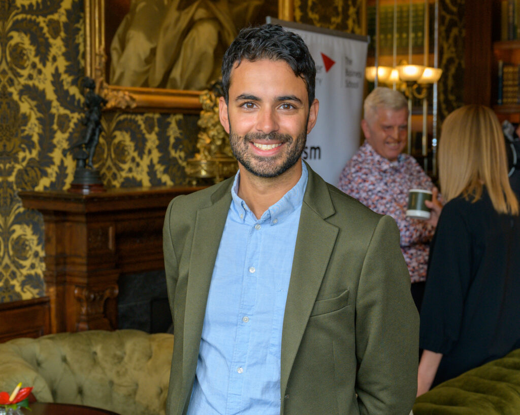 Joshua Ryan-Saha, Director of the Traveltech Innovation Hub at Edinburgh Futures Institute, smiling at camera while wearing olive green blazer over light blue dress shirt at professional networking event in ornate venue with gold decorative wallpaper and chandelier lighting