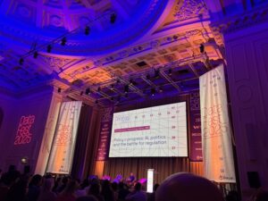 The interior of an ornate venue with elegant architectural details illuminated in purple and blue lighting for DataFest 2025. A large projection screen displays a presentation titled "Policy v progress: AI, politics and the battle for regulation" with a graph showing numeric data points. The DataFest 2025 logo appears in red neon-style text on banners flanking the stage. The audience can be seen in silhouette in the foreground, facing the presentation.