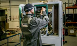 Nucore Group engineer in branded coveralls working on electrical control panel in Aberdeen manufacturing facility, adjusting components inside open cabinet with green interior lighting in industrial workshop setting