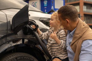 a young child and her father charing up an electric car. The man is wearning a brown gilet with blue shirt and the girl is wearing a striped top. The car is black.