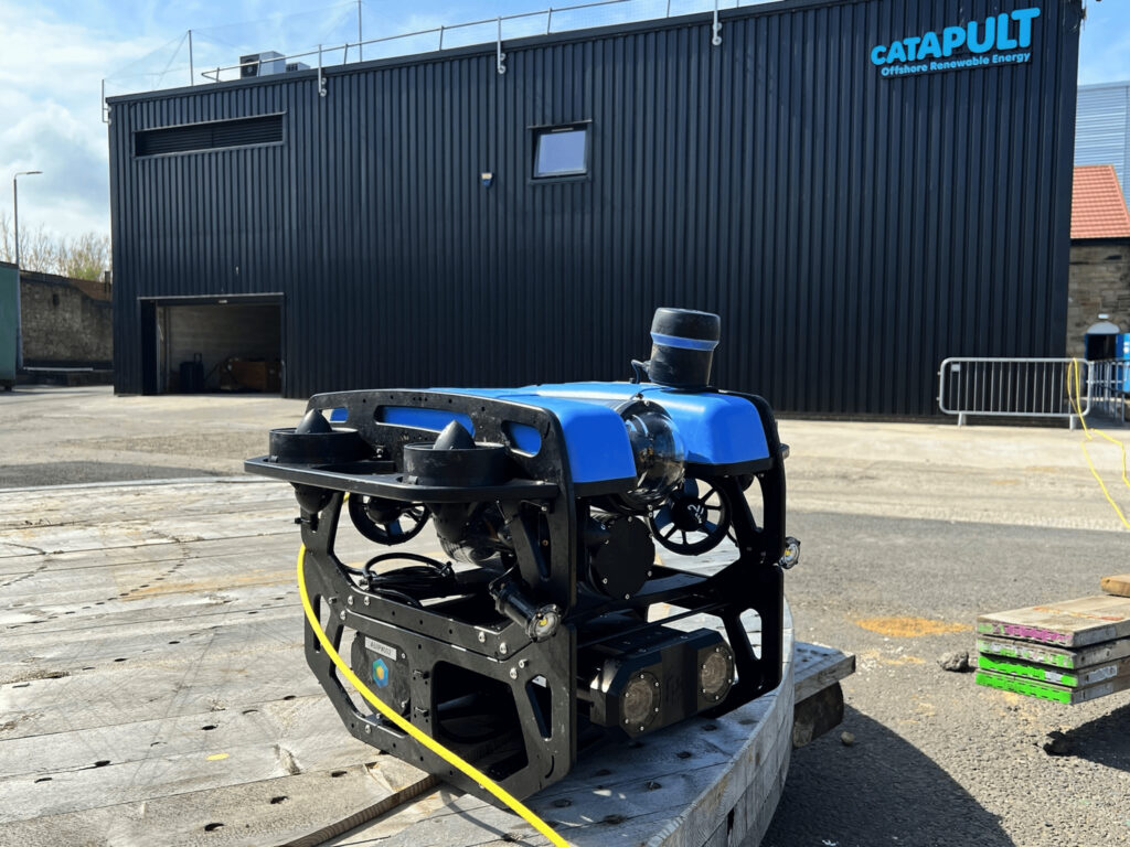 Industrial remotely operated underwater vehicle (ROV) with blue flotation modules, black frame, multiple cameras and propulsion thrusters, connected by yellow tether cable, displayed on concrete testing platform at ORE Catapult's facility with dark corrugated metal building in background