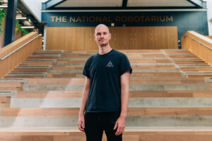 Professional headshot of a man wearing a black t-shirt with a small triangular logo, standing confidently in front of modern wooden amphitheater-style steps at The National Robotarium facility, with the facility name visible on the wall behind him