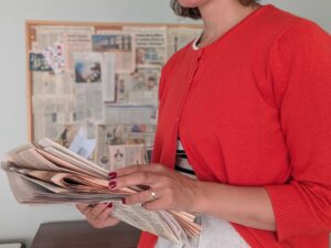 "A woman in a red cardigan with dark nail polish holds a stack of folded newspapers while standing at a desk. Behind her is a cork board covered with newspaper clippings, photos, and various media articles, suggesting a media monitoring or PR workspace environment."