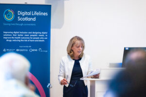 Professor Margaret Whoriskey MBE Head of Innovation for Care Well Being at DHI speaks at a-DLS event. She wear a white jacket and is standing at a lectern with a blue pop up banner nehind her.