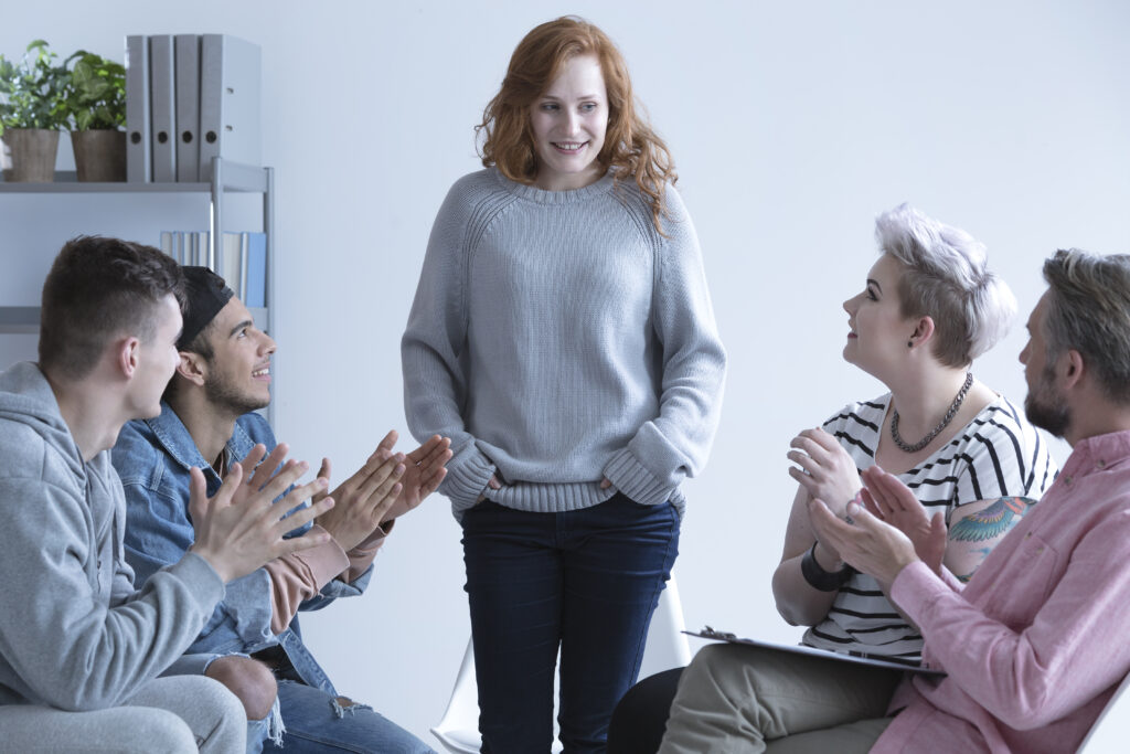 A woman with curly red hair wearing a gray sweater stands in the center of a modern office or meeting room, smiling as four colleagues seated around her applaud. The group appears to be celebrating an achievement or presentation. The setting has white walls, shelving with binders and plants, and the atmosphere suggests a positive team moment or recognition.