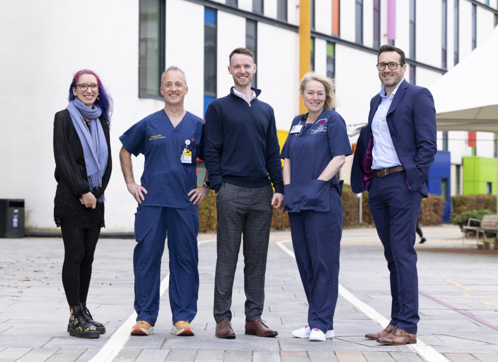 Group photo of five people standing on paved ground outside a modern hospital building with a distinctive white facade featuring colorful window panels in blue, orange, pink, and green. From left to right: a woman with purple hair wearing glasses, black clothing and a light blue scarf; a man in blue NHS medical scrubs; a man in dark casual business attire; a woman in blue medical scrubs; and a man in a dark business suit. The hospital's contemporary architecture with its bright accent colors is visible in the background.