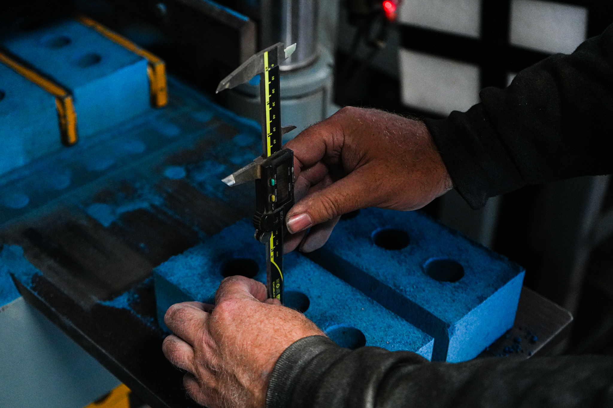Close-up of weathered hands using digital calipers to measure a blue foam block with multiple holes. The person is wearing a dark sweater and appears to be in a workshop or manufacturing setting, with the precision measuring tool positioned to gauge dimensions of the perforated blue material.