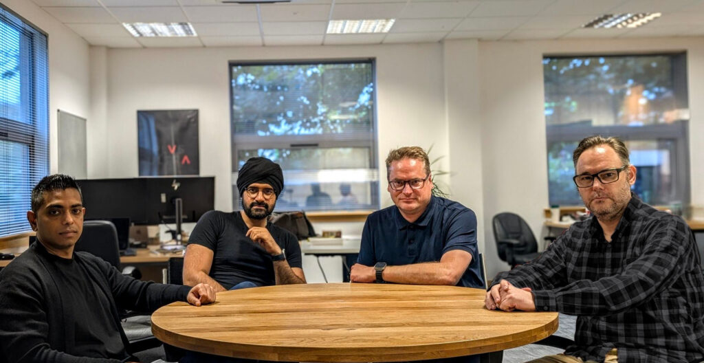 Four men sitting around a circular wooden conference table in a modern office setting. From left to right: a man in a dark sweater, a man wearing a black turban and dark shirt, a man in a navy polo shirt with glasses, and a man in a dark checkered shirt with glasses. The office features large windows with blinds, multiple computer monitors, and contemporary furniture. This appears to be a professional team photo taken in a corporate workspace.