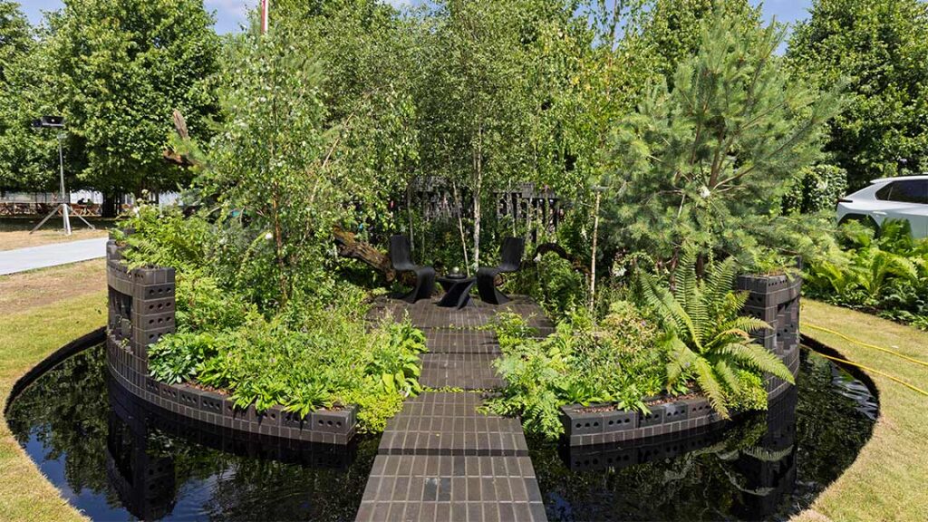 Circular garden installation featuring sustainable K-BRIQ recycled bricks forming curved planters and walkway, surrounded by water feature, showcasing lush ferns and native plants in landscaped outdoor setting with trees and grass background