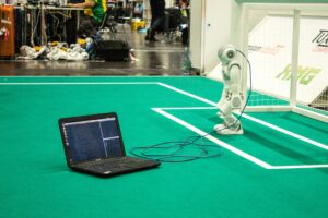 White humanoid robot with camera head connected via cable to laptop computer displaying code on bright green marked floor at robotics developer conference demonstration area