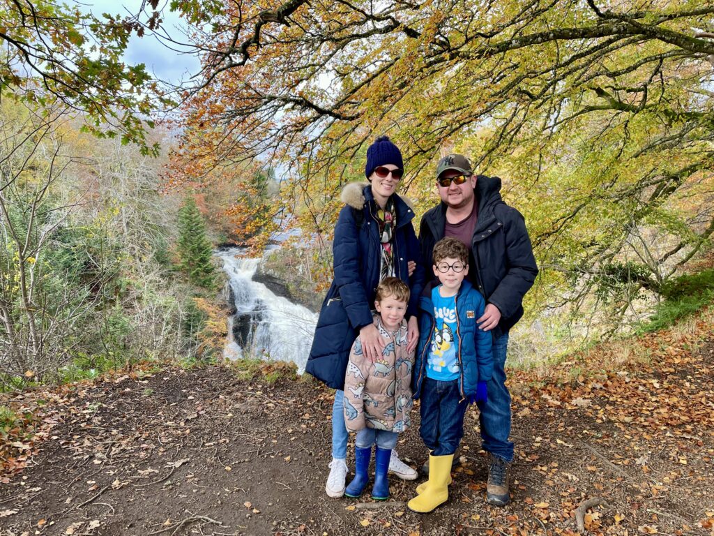 Richard MacQueen, a British Sign Language user featured in the SignPort press release, poses with his family including his partner and two young sons in front of a picturesque waterfall. The family stands on a woodland path surrounded by trees displaying vibrant autumn colors of yellow, orange and green. The scenic Scottish landscape features a multi-tiered waterfall in the background.