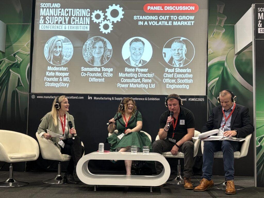 Four panelists seated at a white curved table during the Scotland Manufacturing & Supply Chain Conference panel discussion titled 'Standing Out to Grow in a Volatile Market.' From left to right: moderator Kate Hooper (Founder & MD, StrategyStory) in beige jacket, Samantha Tonge (Co-Founder, B2Be Different) in green dress, Rene Power (Marketing Director/Consultant, Rene Power Marketing Ltd) in black shirt, and Paul Sheerin (Chief Executive Officer, Scottish Engineering) in dark suit. All wear headsets with microphones and red conference lanyards. A large screen behind them displays their names and titles with the conference branding and gear graphics.