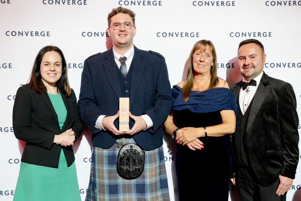 Four people posing at a Converge awards ceremony in front of a branded backdrop. From left to right: a woman in a black blazer and turquoise dress, a young man in a navy suit jacket and tartan kilt holding a wooden award trophy and wearing a sporran, a woman in a navy off-shoulder dress, and a man in a black tuxedo with bow tie. All are smiling at the camera.