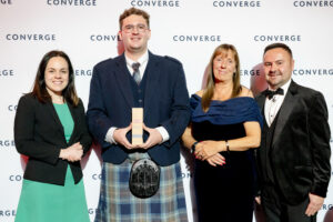 Four people posing at a Converge awards ceremony in front of a branded backdrop. From left to right: a woman in a black blazer and turquoise dress, a young man in a navy suit jacket and tartan kilt holding a wooden award trophy and wearing a sporran, a woman in a navy off-shoulder dress, and a man in a black tuxedo with bow tie. All are smiling at the camera.