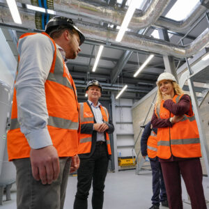 Climate Minister Katie White OBE MP conducts a site visit at the Leeds PIPES Heat Network's Cross Green Energy Centre. The Minister, wearing orange high-visibility safety gear and a white hard hat, is shown in discussion with three representatives from Vital Energi, all wearing similar safety equipment. They stand inside the energy centre surrounded by large industrial heat exchange equipment, insulated pipes, and thermal infrastructure. The facility's high ceiling and industrial architecture are visible in the background.