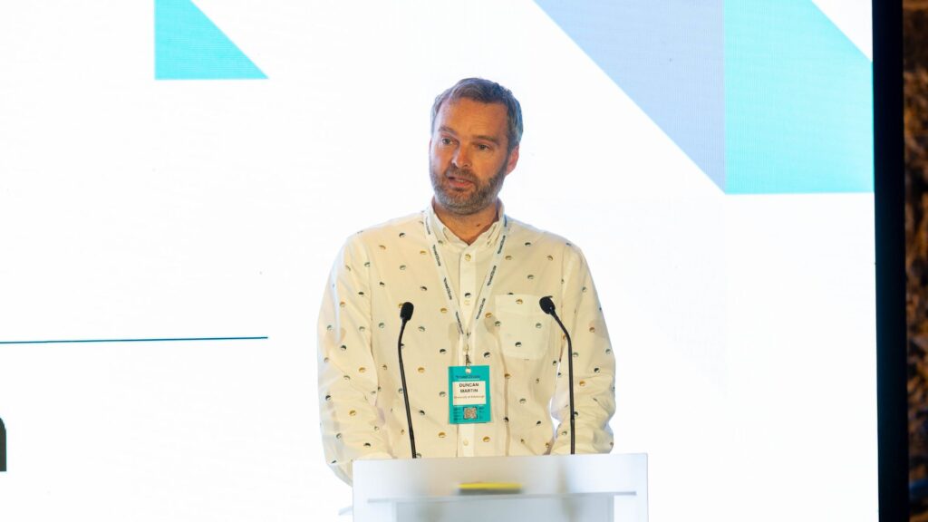 Male speaker wearing a cream-colored shirt with small pattern standing at a transparent podium with two microphones, presenting at the Invest2Scale conference. Modern geometric backdrop features light blue and turquoise angular shapes. Partial text 'entre' visible on left side. Teal armchair with blue Techscaler cushion in foreground, audience members visible at bottom of frame.