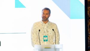 Male speaker wearing a cream-colored shirt with small pattern standing at a transparent podium with two microphones, presenting at the Invest2Scale conference. Modern geometric backdrop features light blue and turquoise angular shapes. Partial text 'entre' visible on left side. Teal armchair with blue Techscaler cushion in foreground, audience members visible at bottom of frame.