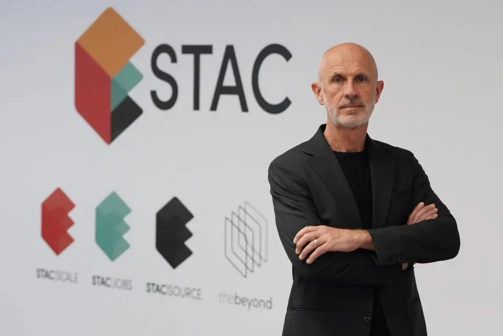 Jim Rowan, Chair of STAC, standing with arms crossed in front of the STAC brand wall, wearing a dark blazer over a black shirt in a professional headshot pose.