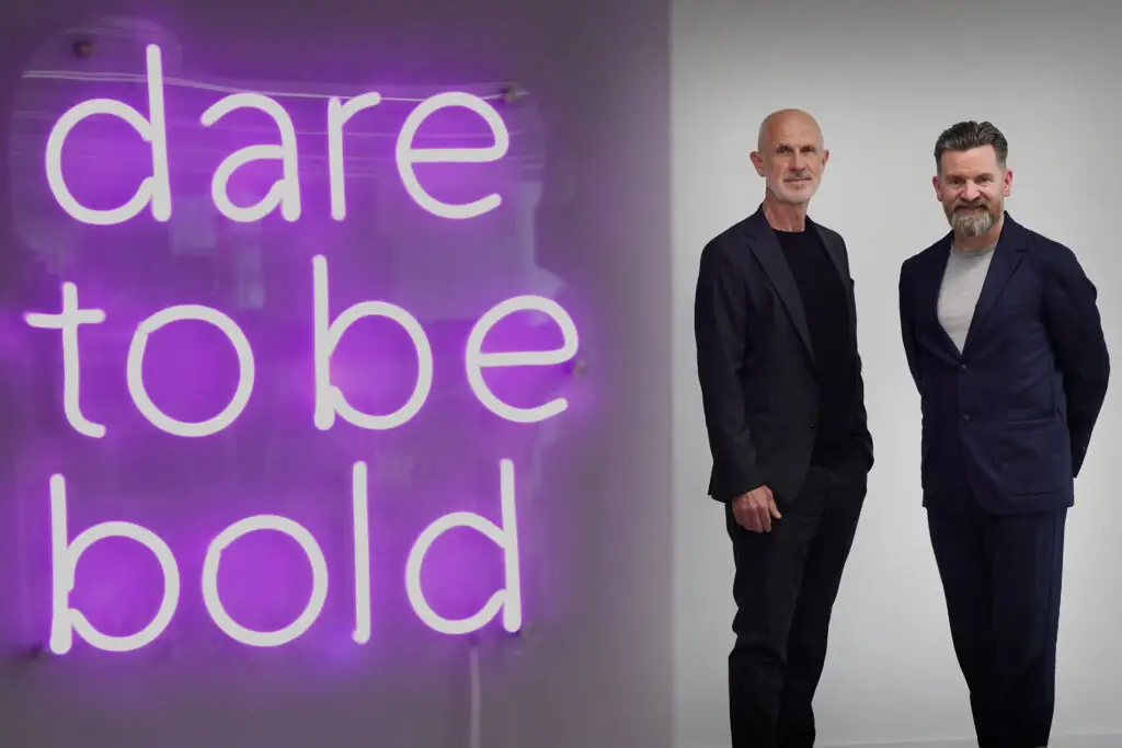 Jim Rowan and Paul Wilson standing in front of a purple neon sign reading 'dare to be bold'. Both men wear dark blazers and are positioned in a professional office setting.