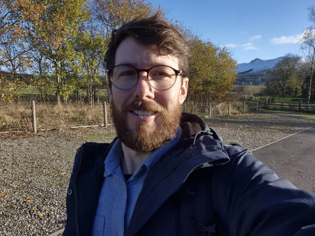 Tristan, 41, from Edinburgh, smiling in an outdoor selfie taken in an autumnal Scottish landscape with a snow-capped hill visible in the background.