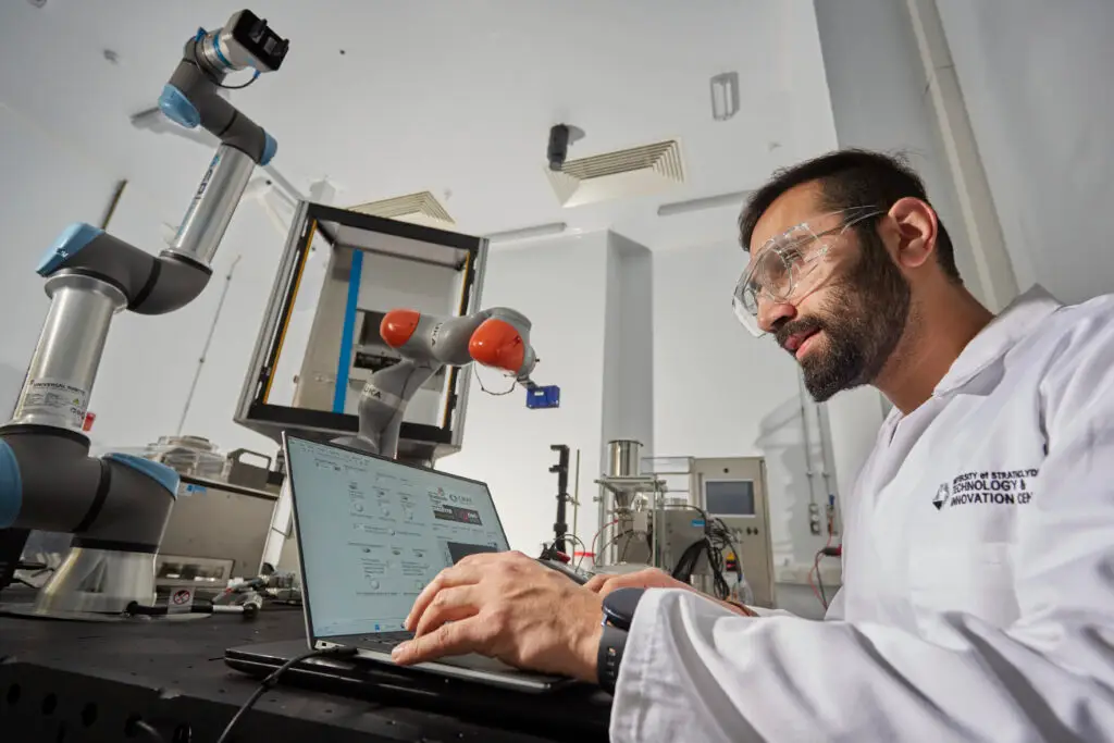 A bearded male researcher wearing a white lab coat and clear safety goggles types on a laptop in a high-tech laboratory, with two robotic arms — a Universal Robots arm and a KUKA arm — visible in the background alongside pharmaceutical manufacturing equipment. The lab coat bears the University of Strathclyde Technology and Innovation Centre logo.