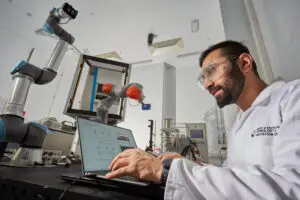 A bearded male researcher wearing a white lab coat and clear safety goggles types on a laptop in a high-tech laboratory, with two robotic arms — a Universal Robots arm and a KUKA arm — visible in the background alongside pharmaceutical manufacturing equipment. The lab coat bears the University of Strathclyde Technology and Innovation Centre logo.