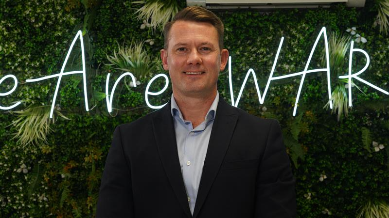 Headshot of Justin Meyer, Managing Director at Evolt Charging, wearing a dark blazer and light blue shirt, standing in front of a green plant wall with a neon sign partially reading "We Are WAR…" in the background.