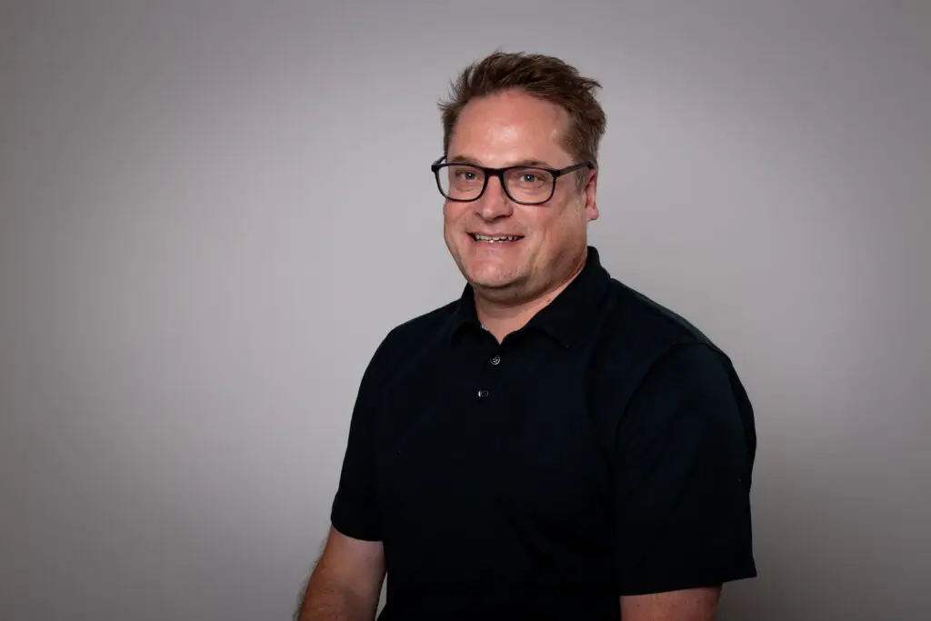 A smiling man with short auburn hair and dark rectangular-framed glasses, wearing a black polo shirt, photographed against a plain light grey background.