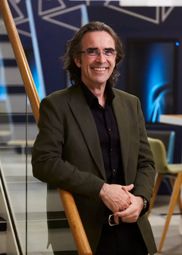 A smiling man with shoulder-length grey-brown wavy hair and rimless glasses, wearing a dark green blazer over a black shirt, leaning against a wooden staircase railing in a modern office space with blue-toned decor in the background.