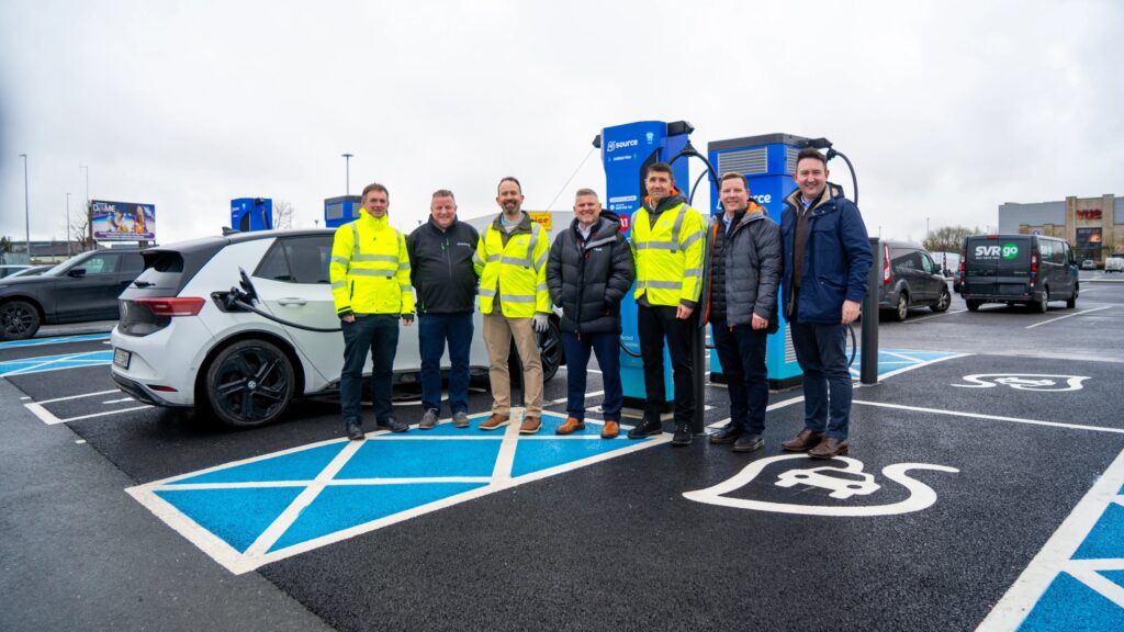 Group photo of seven representatives from Source, Siemens eMobility, and Evolt Charging standing beside a Source-branded EV charging unit at Ashbourne Retail Park. Several wear high-visibility yellow jackets. A white Volkswagen electric vehicle is plugged in to the left, and EV charging bay markings are visible on the ground.