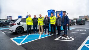 Group photo of seven representatives from Source, Siemens eMobility, and Evolt Charging standing beside a Source-branded EV charging unit at Ashbourne Retail Park. Several wear high-visibility yellow jackets. A white Volkswagen electric vehicle is plugged in to the left, and EV charging bay markings are visible on the ground.
