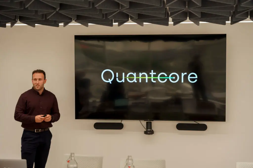 Jack Brennan, co-founder and CEO of Quantcore Technologies, presents in front of a large screen displaying the Quantcore logo at a pitch or investor showcase event in Glasgow.