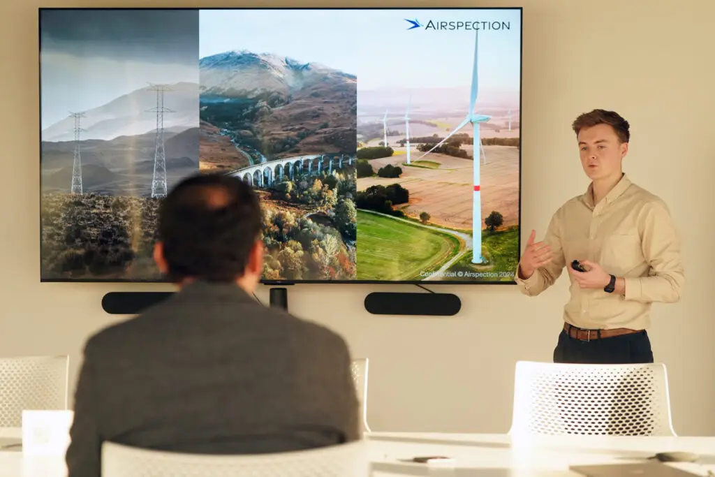 Matthew McLean, founder and Director of Airspection, presents to a seated audience in front of a large screen showing the Airspection brand and imagery of offshore wind turbines, power lines and Scottish Highland landscapes, illustrating the company's infrastructure inspection technology.