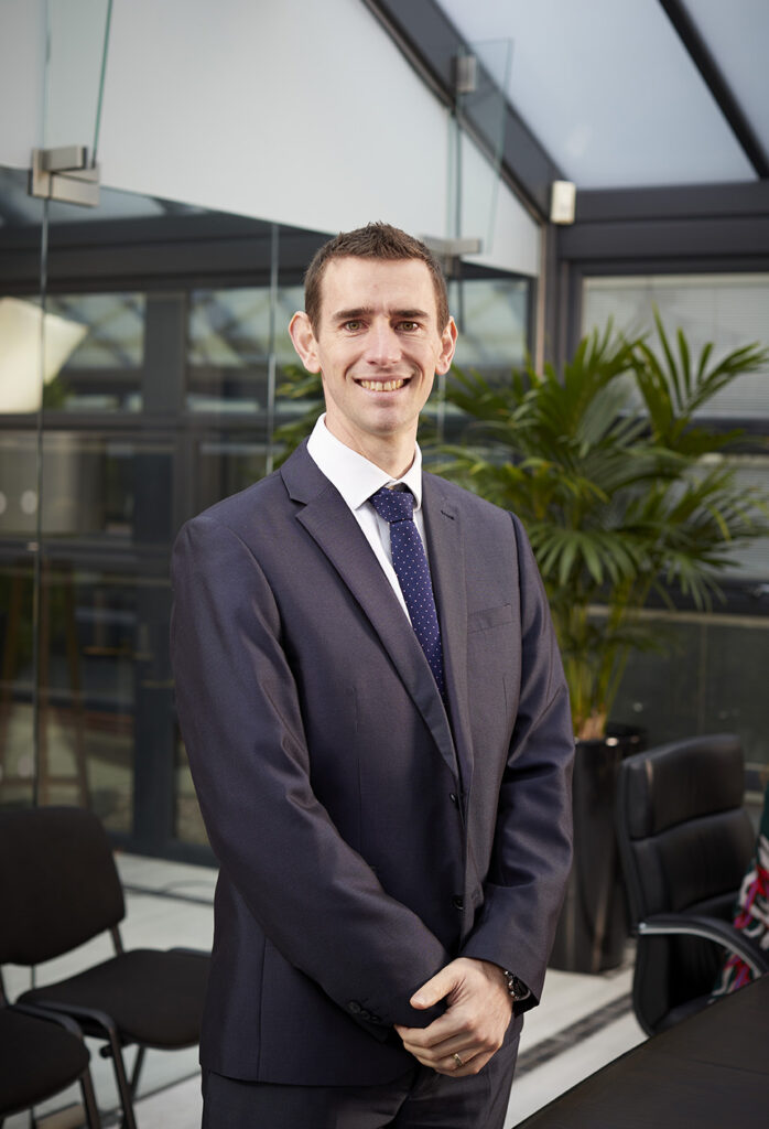 John Runniff, Account Development Director at Vital Energi, smiling and standing in a modern office atrium wearing a dark suit and navy tie.