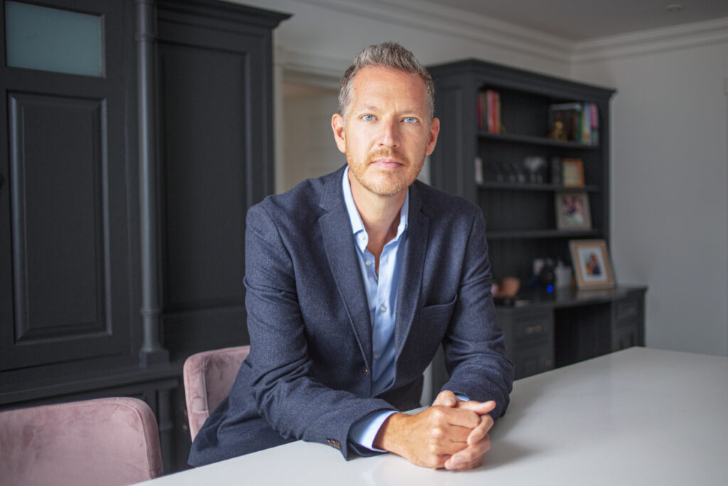 Nick Thompson, Co-Founder and CEO of OLO Robotics, photographed seated at a table in a dark-furnished interior setting, speaking to the commercial launch of OLO Robotics and its browser-based ROS2 platform bringing robotics development to mainstream software teams in warehouses, factories and labs