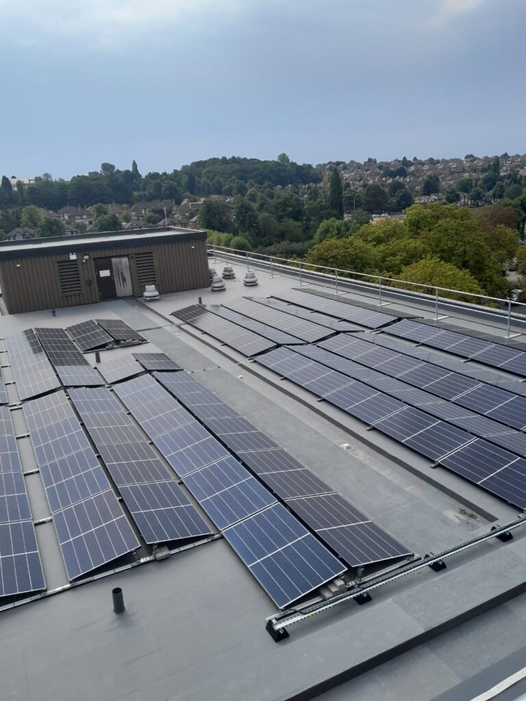 Rows of solar panels installed across the flat roof of Nottingham City Hospital, with green hillside housing visible in the background under a clear blue sky.