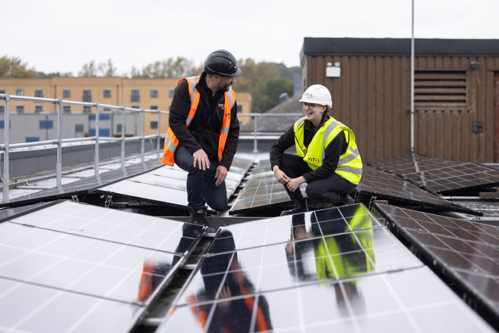 Two Vital Energi engineers in hi-vis vests and hard hats crouching on a hospital rooftop, inspecting newly installed solar panels and smiling.