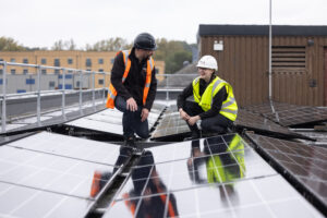 Two Vital Energi engineers in hi-vis vests and hard hats crouching on a hospital rooftop, inspecting newly installed solar panels and smiling.