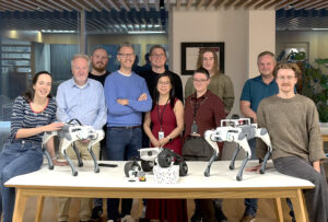 The OLO Robotics team photographed together in an office setting, left to right Lenka Mudrich, Stephan Holingshead, George Bridges, Nick Thompson, Simon I Anson, Eleanor Tang-Smith, Danielle Clayton, Sam Piper, Matthew Abdy and Sam Gibson, with two quadruped robots and a wheeled mobile robot on the table in front of them, marking the Sheffield startup's commercial launch in May 2026
